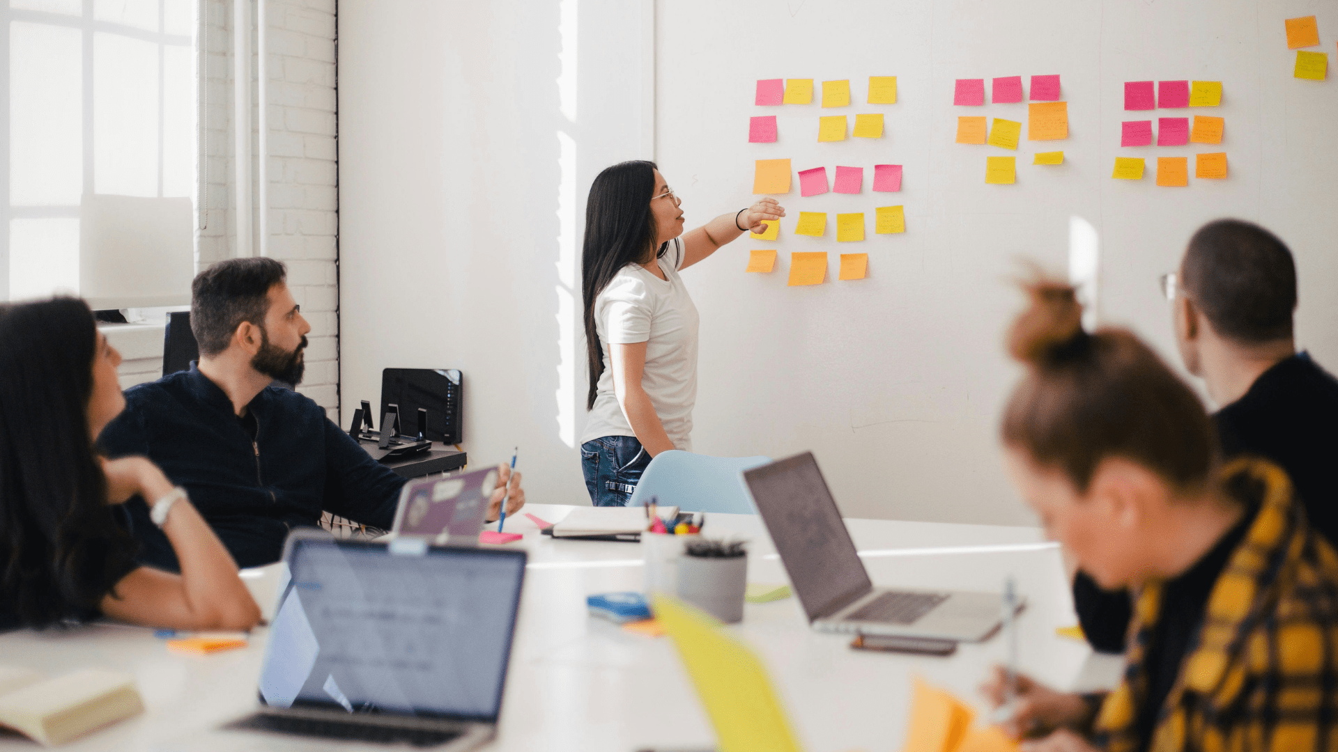 Pessoas sentadas ao redor de uma mesa, em que à frente de todas há uma mulher apontando para um quadro branco no qual estão fixados post-its