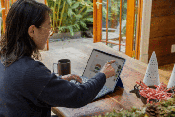 Mulher sentada à mesa usando um tablet com teclado, apontando para a tela com a mão direita. Ao lado há uma caneca preta e uma tigela com bengalas de doce, sobre uma mesa de madeira, com porta de vidro e plantas ao fundo.