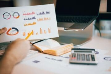 Folhas com gráficos, calculadora, caderno e notebook se encontram sobre uma mesa. Uma mão segura um lápis e uma folha de papel, na qual se encontram números, gráficos coloridos e porcentagem financeira.
