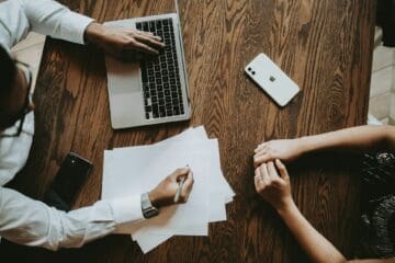Governança corporativa: fotografia mostra homem e mulher sentados à mesa, sendo que o primeiro está segurando uma caneta e anotando informações em um papel, enquanto digita com a outra mão no teclado de um notebook; já a segunda aparece somente com as mãos sobre a mesa