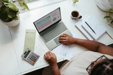 Sobre uma mesa se encontram: notebook, calculadora, planilhas, cadernos e xícara com café