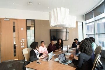 Um grupo de oito pessoas está reunido em uma sala de conferências moderna, sentadas ao redor de uma mesa retangular de madeira. Todas estão voltadas para a discussão, com alguns notebooks abertos sobre a mesa. Há uma grande luminária branca no teto e uma televisão desligada presa à parede ao fundo. A sala possui janelas amplas com cortinas cinzas que permitem a entrada de luz natural.