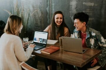 Três pessoas sentadas em uma mesa de madeira, sorrindo e conversando enquanto trabalham em laptops, em um ambiente descontraído de escritório ou café.