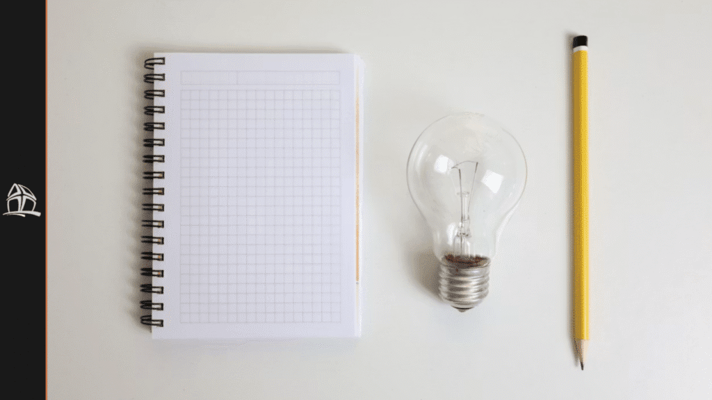 Caderno, lâmpada e lápis sobre a mesa. Pessoa navega em seu notebook, sobreposto à mesa. Trabalho remoto, híbrido ou presencial.