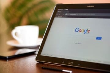 Um tablet Samsung sobre uma mesa de madeira exibindo a página inicial de buscas do Google. Ao fundo, uma xícara de café desfocada.
