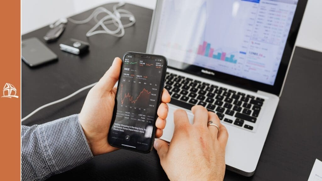 Uma pessoa segurando um smartphone que mostra um gráfico de linha de desempenho financeiro (Yahoo Finance), enquanto a outra mão está sobre o teclado de um laptop que também exibe gráficos.
