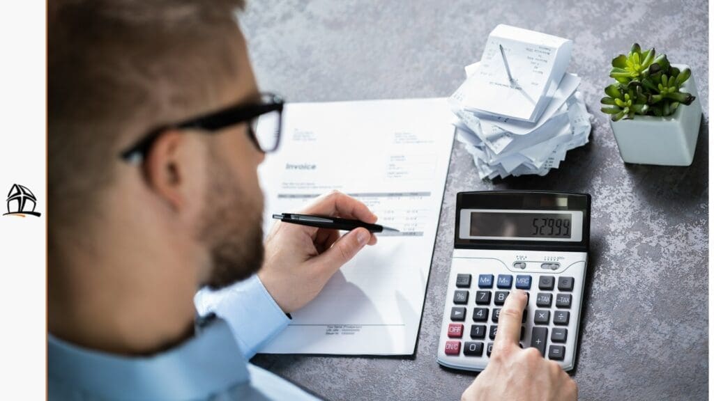 Visão por cima do ombro de um homem usando óculos enquanto faz contas em uma calculadora de mesa ao lado de uma fatura e uma pilha de recibos presos em um espeto.